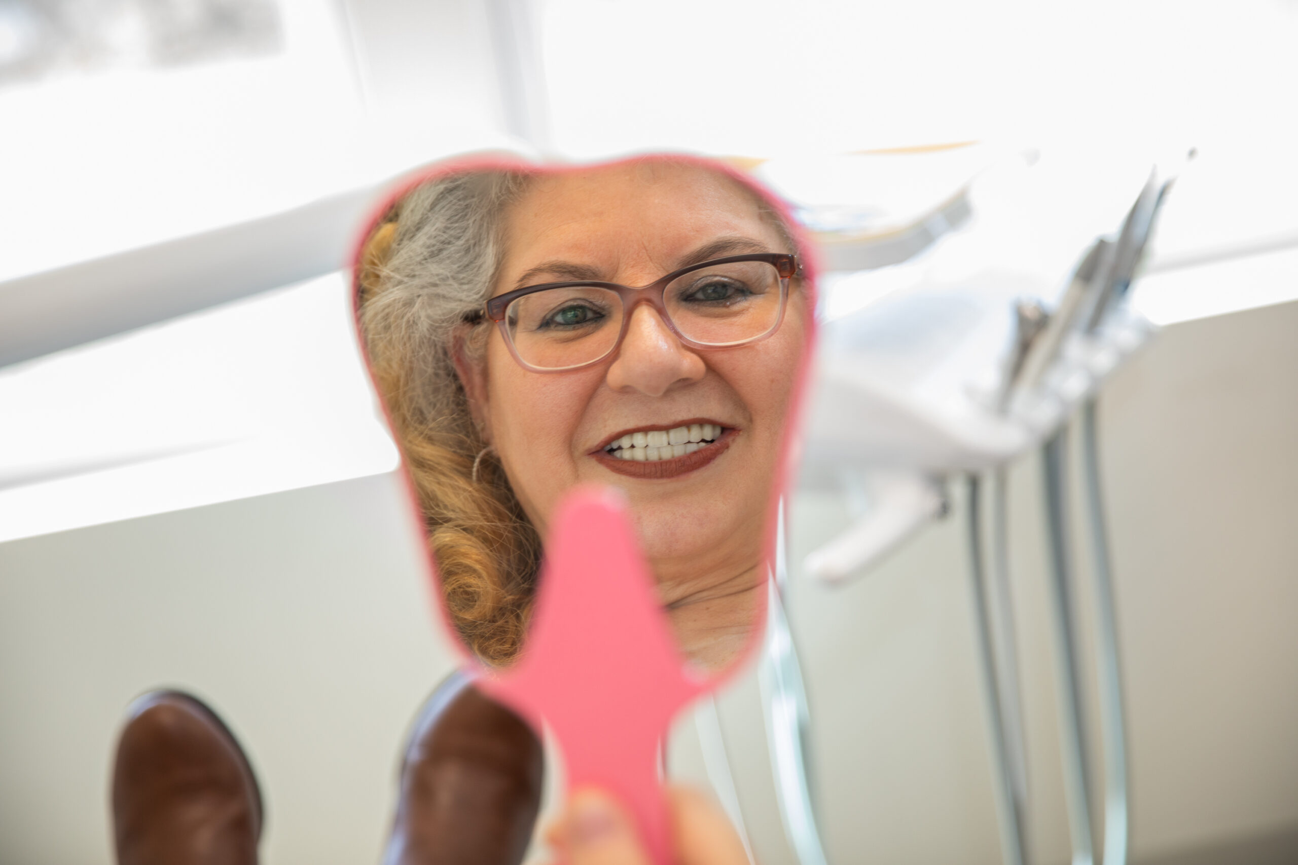 a patient smiling at her new teeth through a pink hand-held mirror after she has been treated with full mouth dental implants in Dr. Martirossian's office.